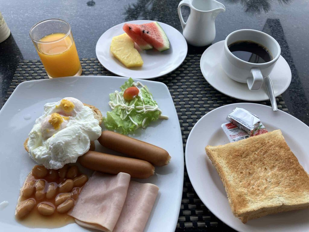 ポーチドエッグやトースト、ソーセージ、ハム、豆、サラダ、フルーツ、コーヒー、オレンジジュースが並ぶ朝食セット